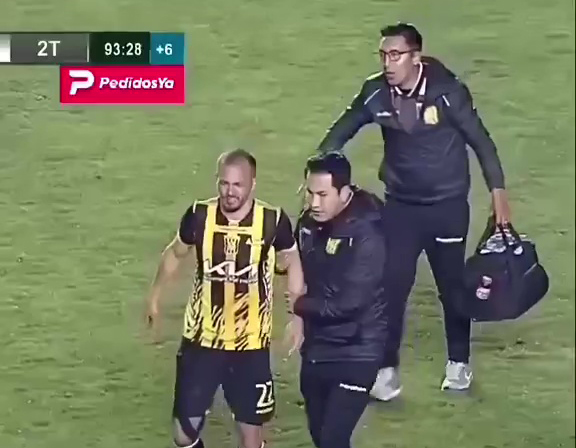 Football player collapsing on the field, assisted by two men.