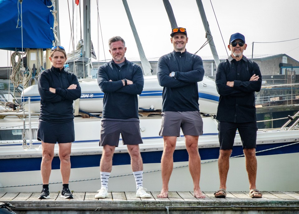 Four people standing on a dock, arms crossed, in front of sailboats.