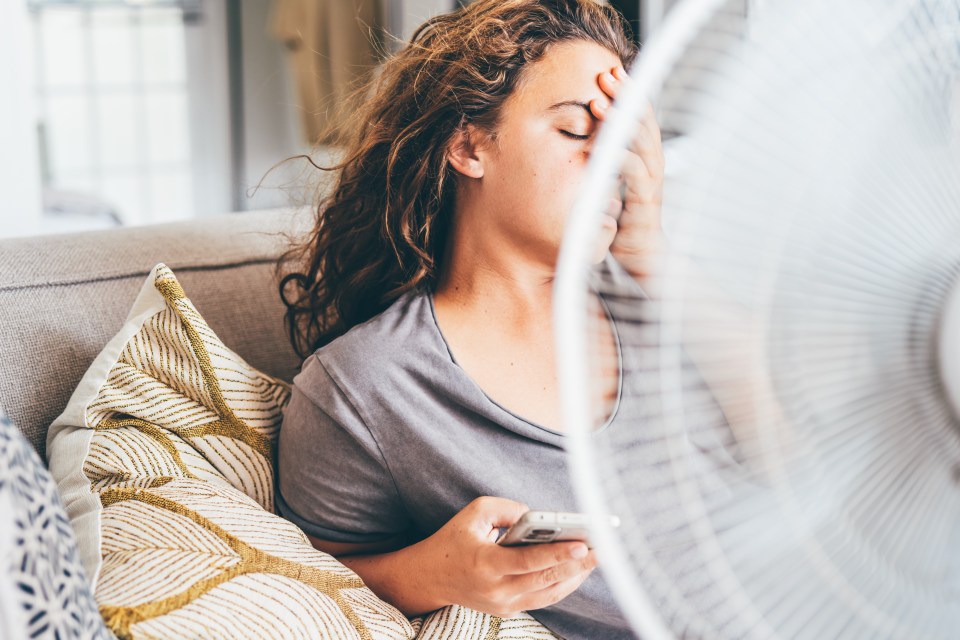 Woman sitting on a couch using an electric fan on a hot day.