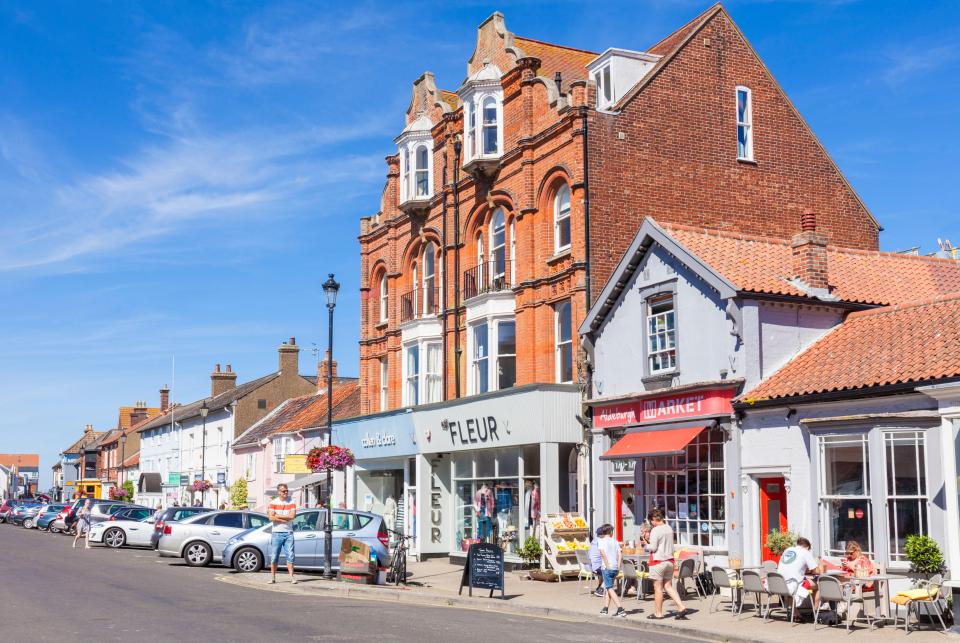 Aldeburgh High Street in Suffolk, England, showing shops and cafes with people browsing and sitting outside.