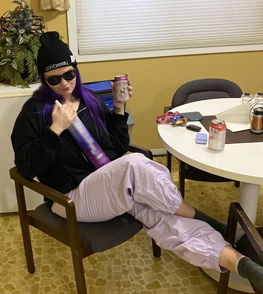 A woman with purple hair sits in a chair, holding a can of black cherry Waterloo sparkling water.