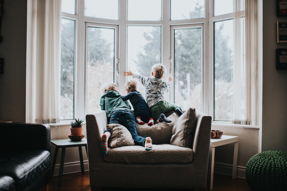 Three children look out a window at falling snow.