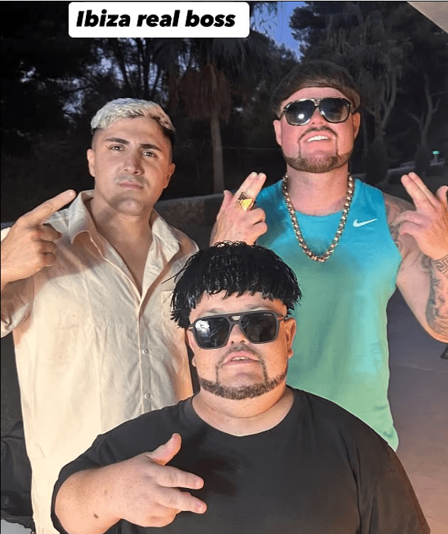 Three men posing, one in the center wearing sunglasses and a dark wig.