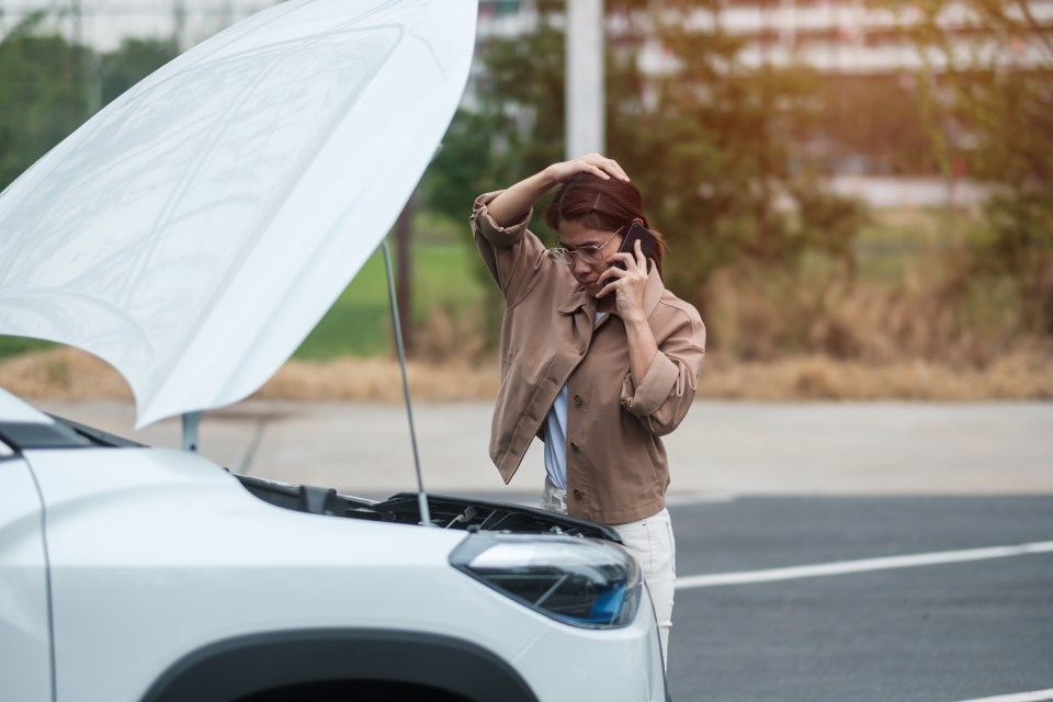 Woman on roadside phone call, car hood open.