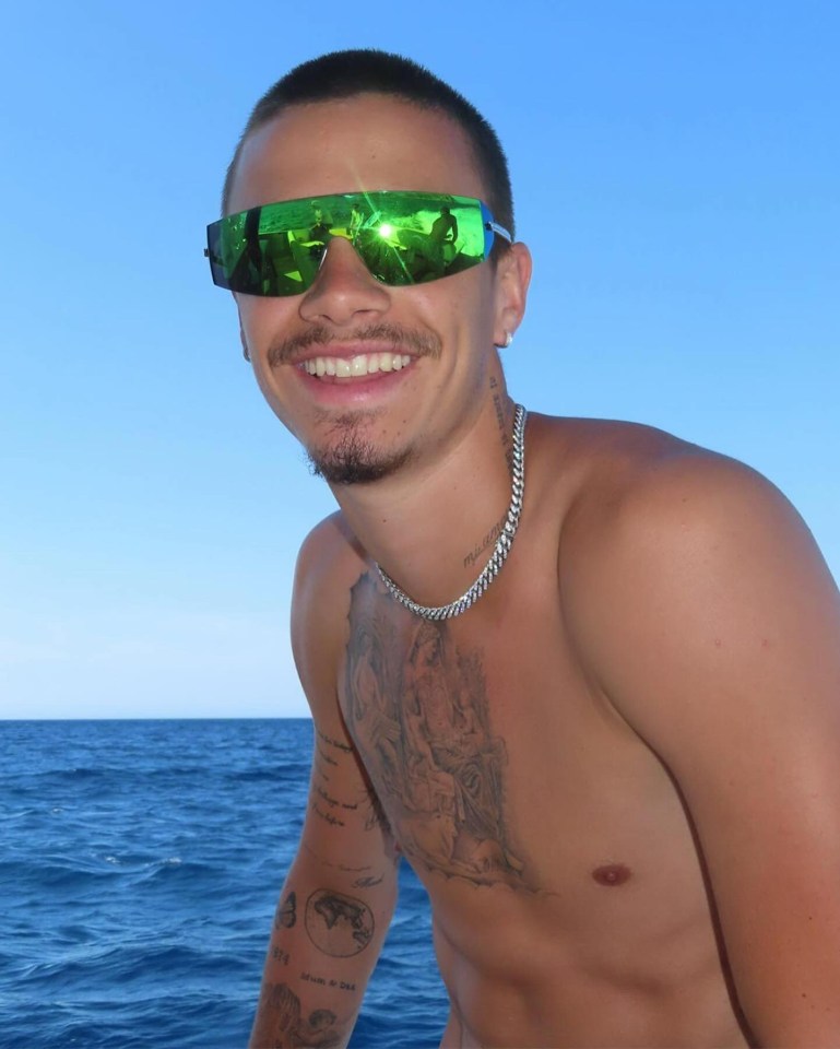 Romeo Beckham smiling, wearing green sunglasses and a silver chain, on a boat at sea.