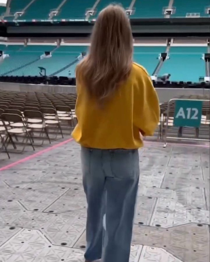 Woman in yellow sweater and jeans walking away from camera in an empty stadium.