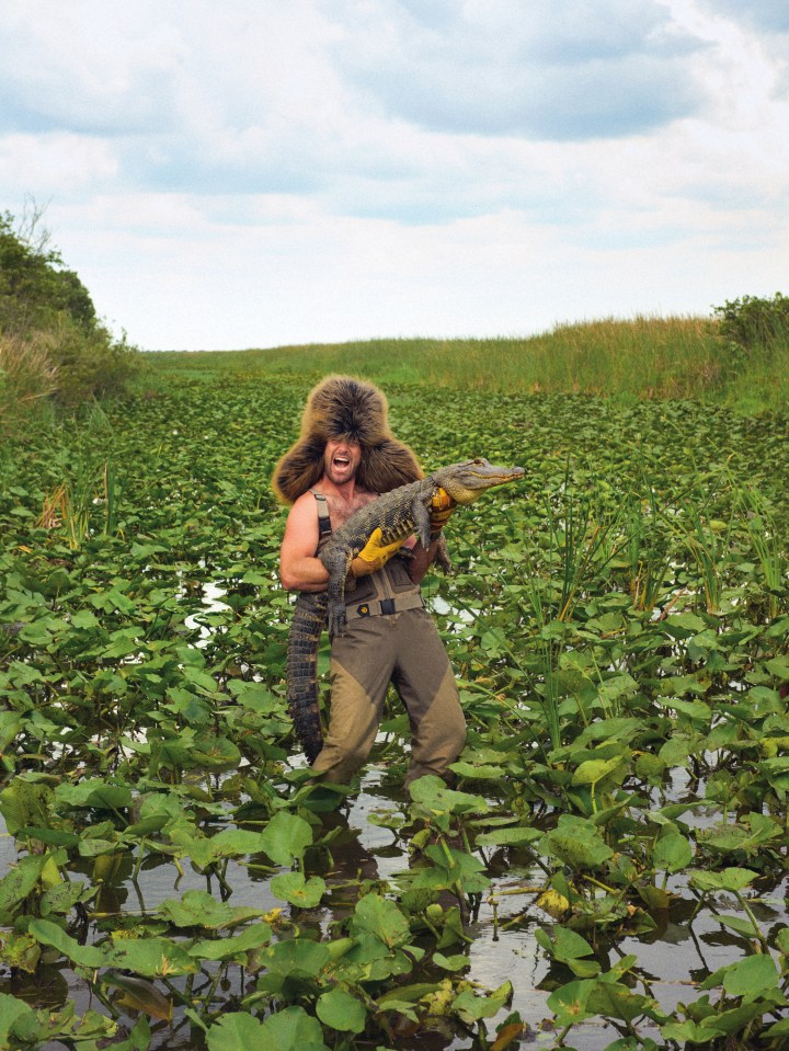 A man in a fur hat and waders holds a large alligator in a swamp.