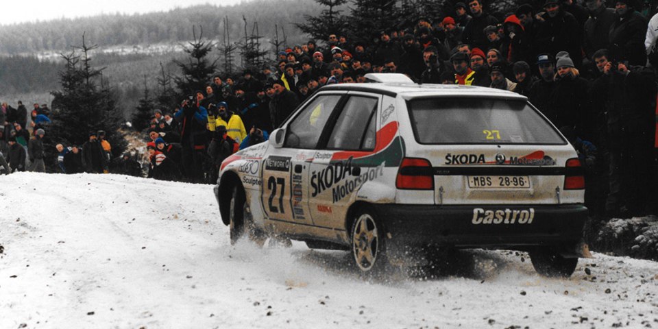 A Škoda rally car driving through snow, watched by spectators.