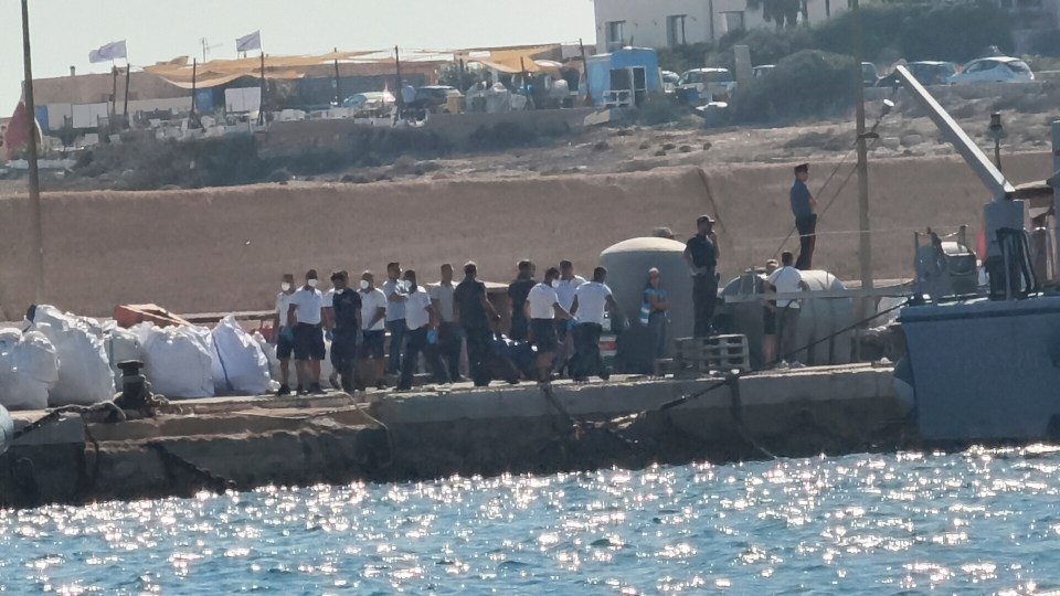 Italian Coast Guard officers carrying a body bag on a dock.