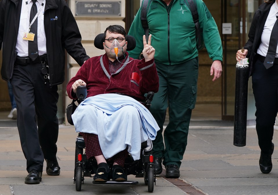 Nicholas Rossi, in a wheelchair and wearing an oxygen mask, gives a peace sign as he leaves court.