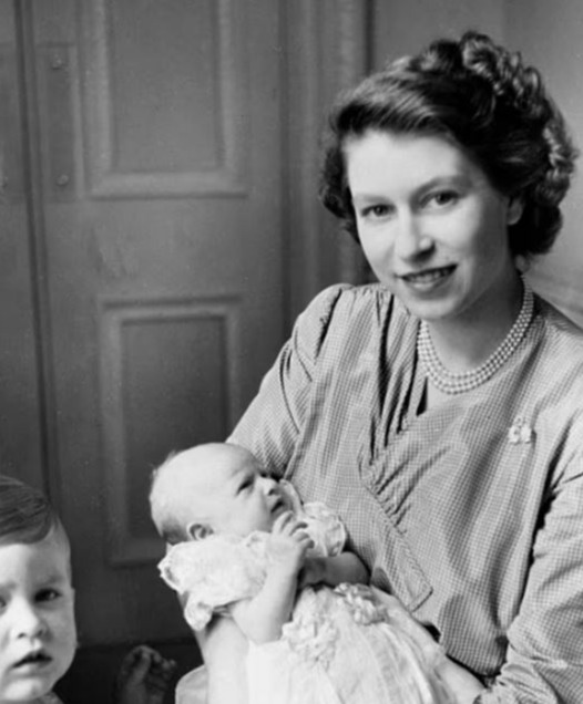 Black and white photo of Princess Anne as a baby, held by her mother, with a young boy beside them.