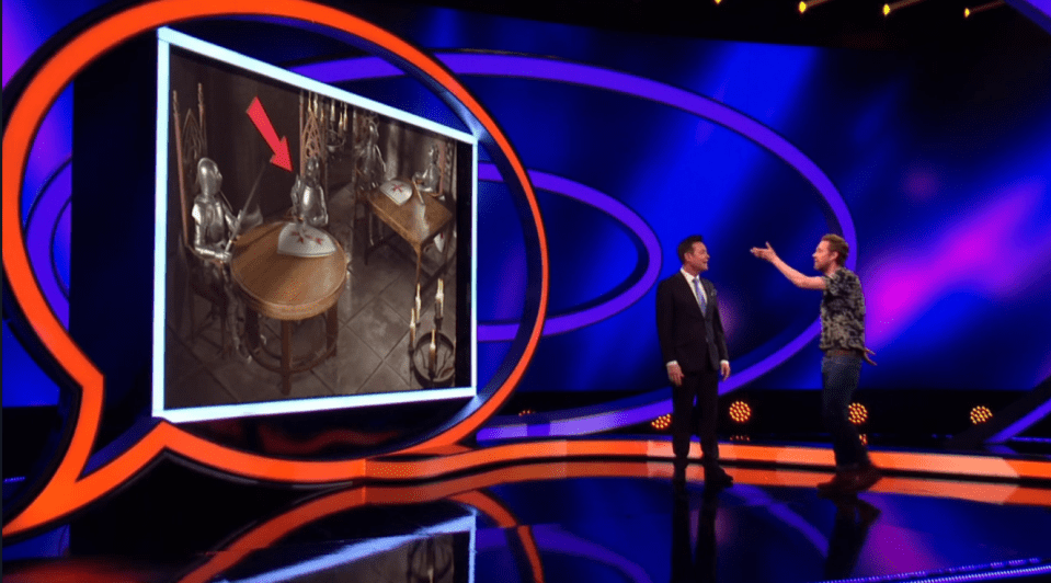 Screenshot of two men on a game show stage, one pointing at a screen showing two knights seated at a table.