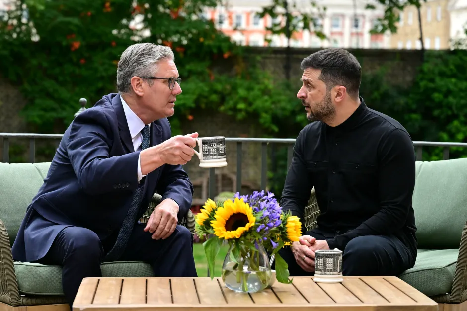 Two men in suits sit outdoors, talking and holding mugs.