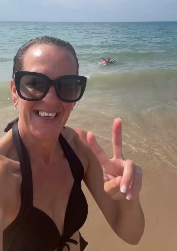 Woman in a bikini on a beach giving a peace sign; a child is bodyboarding in the background.