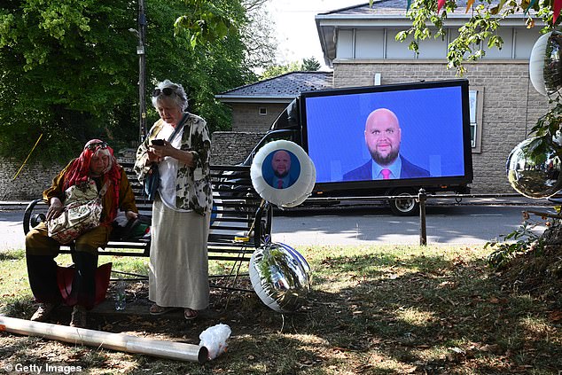 Two women rest on a bench surrounded by JD Vance balloons as a van displaying the Vance mem drives past them through Charlbury