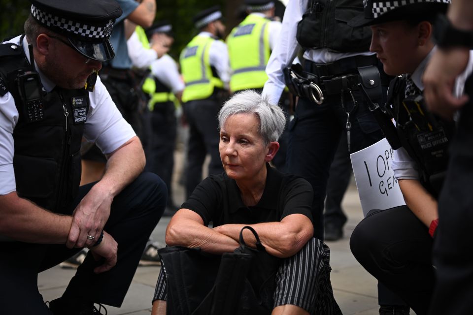 A woman sits with arms crossed as two police officers kneel beside her at a protest.