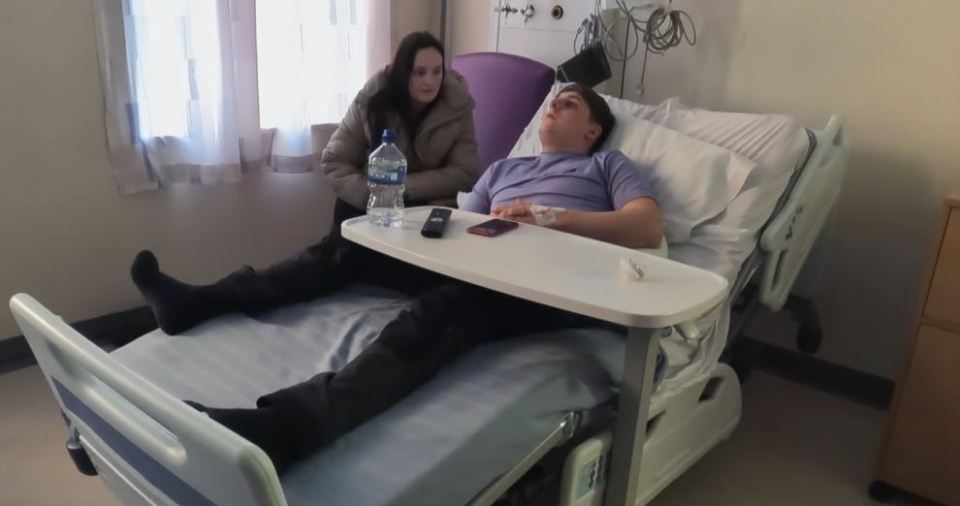 A man lies in a hospital bed with a woman sitting beside him.