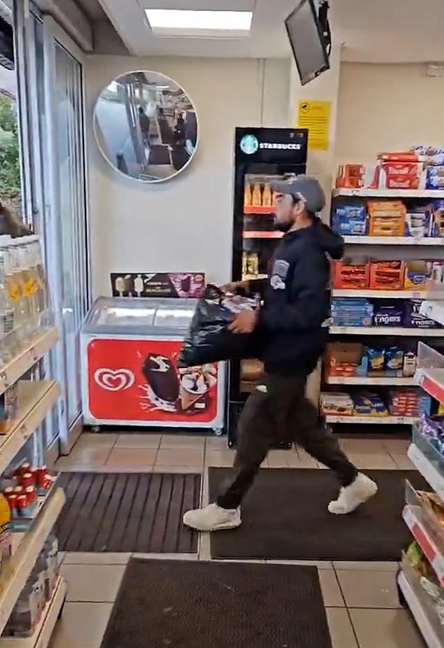 The man is not alone in his efforts - an accomplice (pictured) soon appears on screen, also carrying a loaded black carrier bag, seemingly having targeted the back of the shop