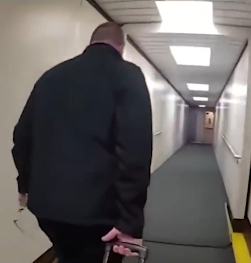 A man in a dark jacket is escorted down an airport hallway.