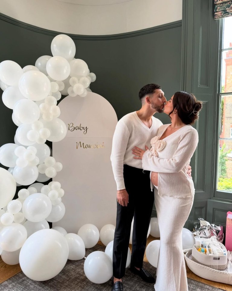 Pregnant couple kissing at baby shower with white balloons and sign saying "Baby Mancini".