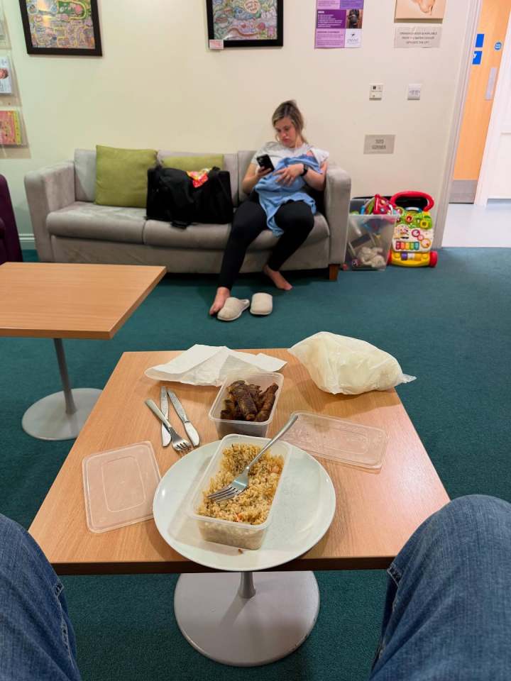 Takeout food on a table in a waiting room; a woman breastfeeds a baby on a nearby couch.