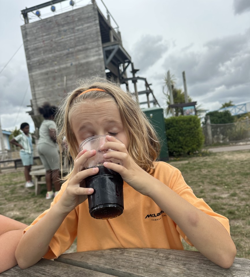 Child drinking from a cup outdoors near a climbing wall.
