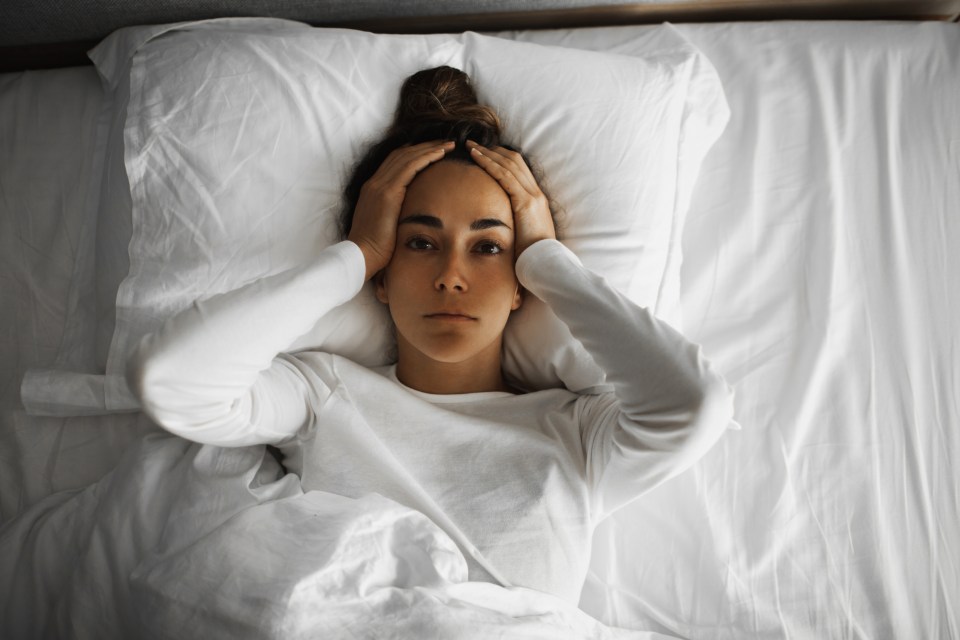 Woman lying in bed, hands on head, looking distressed.
