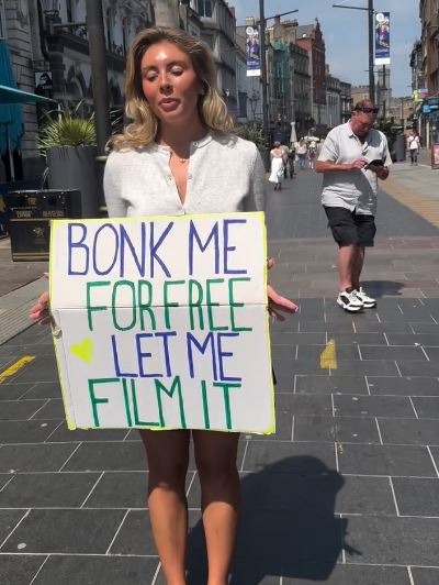 Woman holding a sign that says "Bonk me for free let me film it" on a city street.