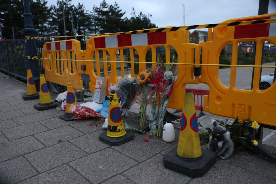 Floral tributes and stuffed animals left at a crash site behind yellow barriers.