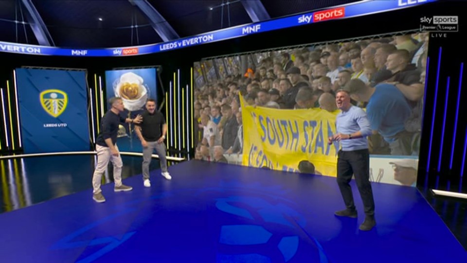 Gary Neville on a Sky Sports set, reacting to a Leeds United fan banner.