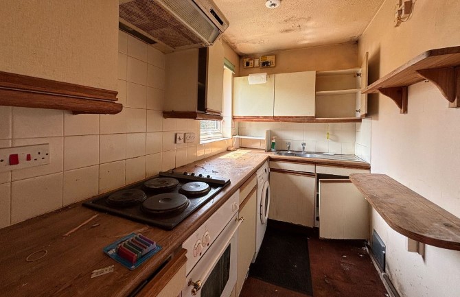 A dilapidated kitchen with wood countertops, off-white cabinets, and a four-burner electric stovetop.