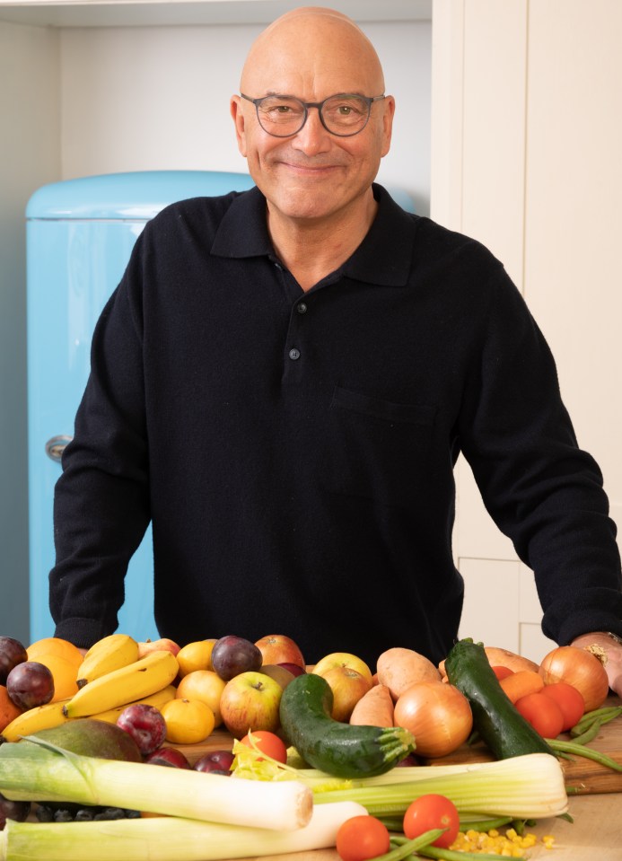 Gregg Wallace leaning on a kitchen counter with a large assortment of fruits and vegetables.
