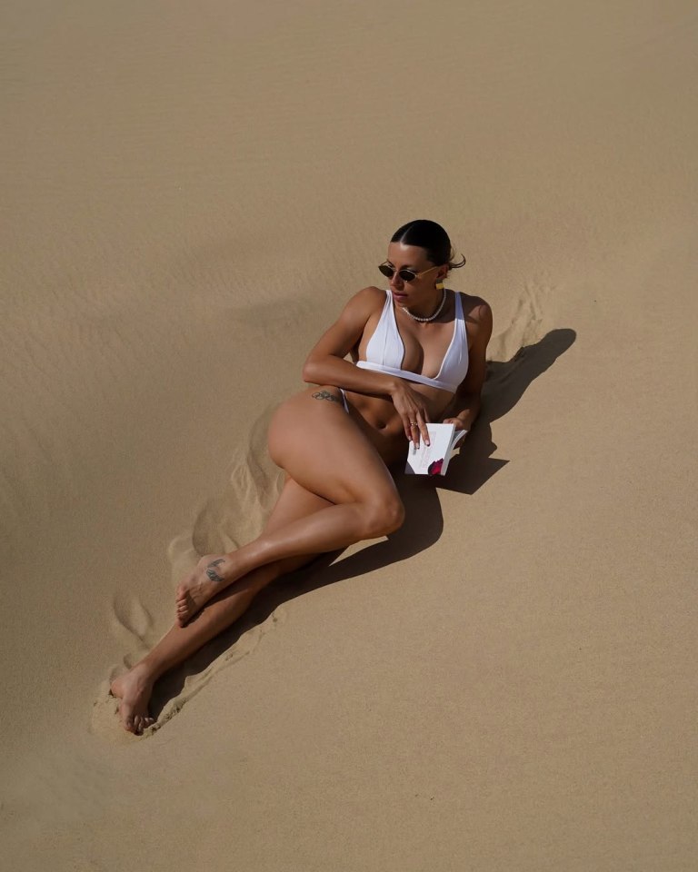 Woman in white bikini lying on sand, reading a book.