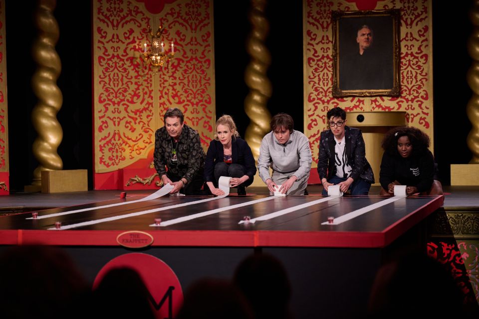 Five contestants unrolling toilet paper on a game show set.