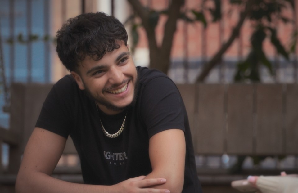 A smiling young man sits outdoors, talking to someone out of focus.