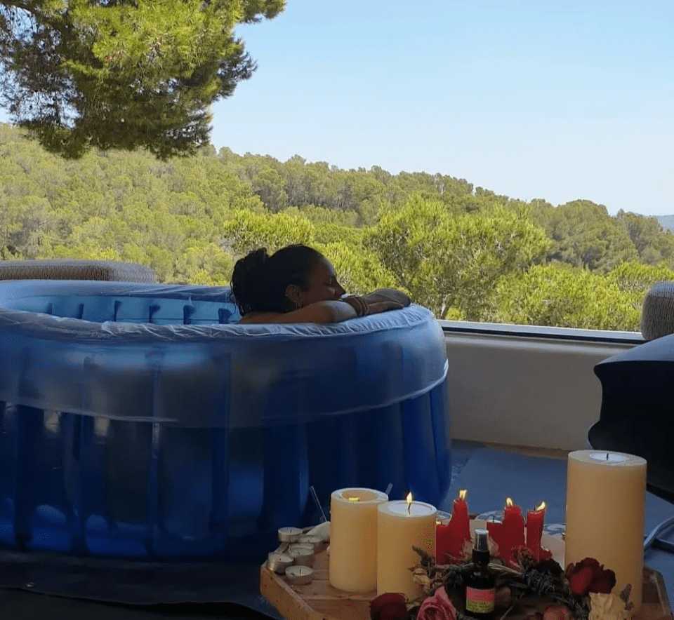 Person relaxing in a blue inflatable pool with candles and flowers nearby, overlooking a hillside.