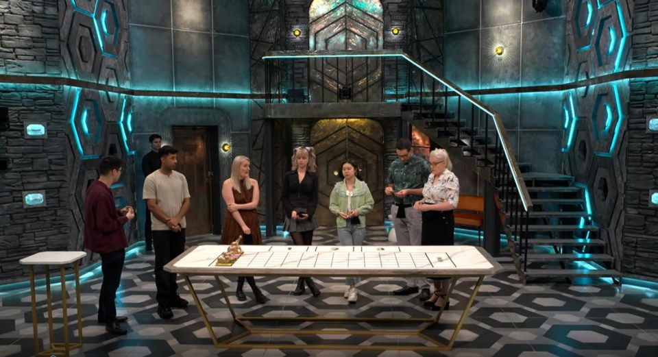 Seven contestants playing a game around a large table in a futuristic-looking set.