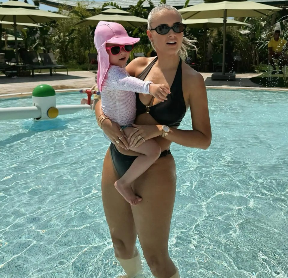 Woman in a black bikini holding a toddler in a pink hat in a pool.