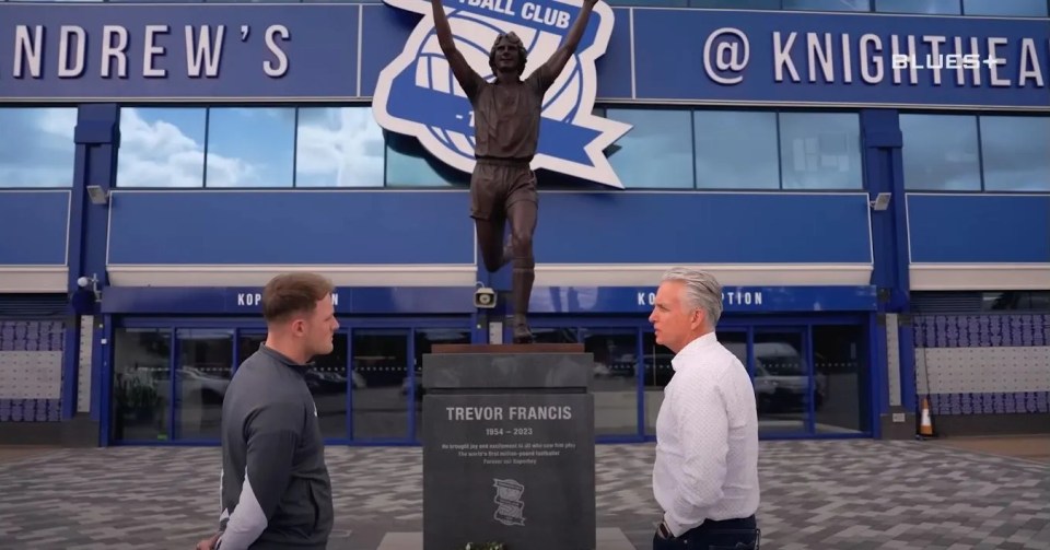 Two men standing by a statue of Trevor Francis at a football stadium.