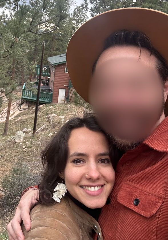 Close-up selfie of a woman with a man in the background; a cabin is visible in the far background.