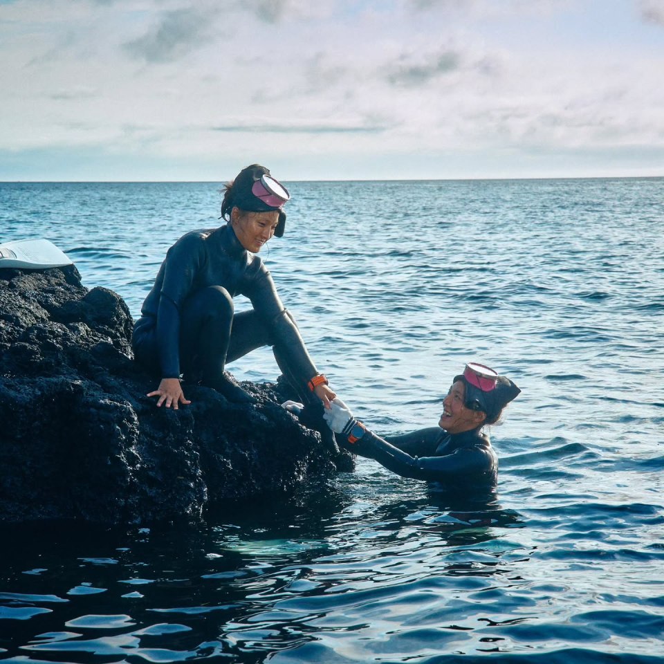 Two Haenyo women, traditional Jeju Island divers, assisting each other near rocks in the ocean.