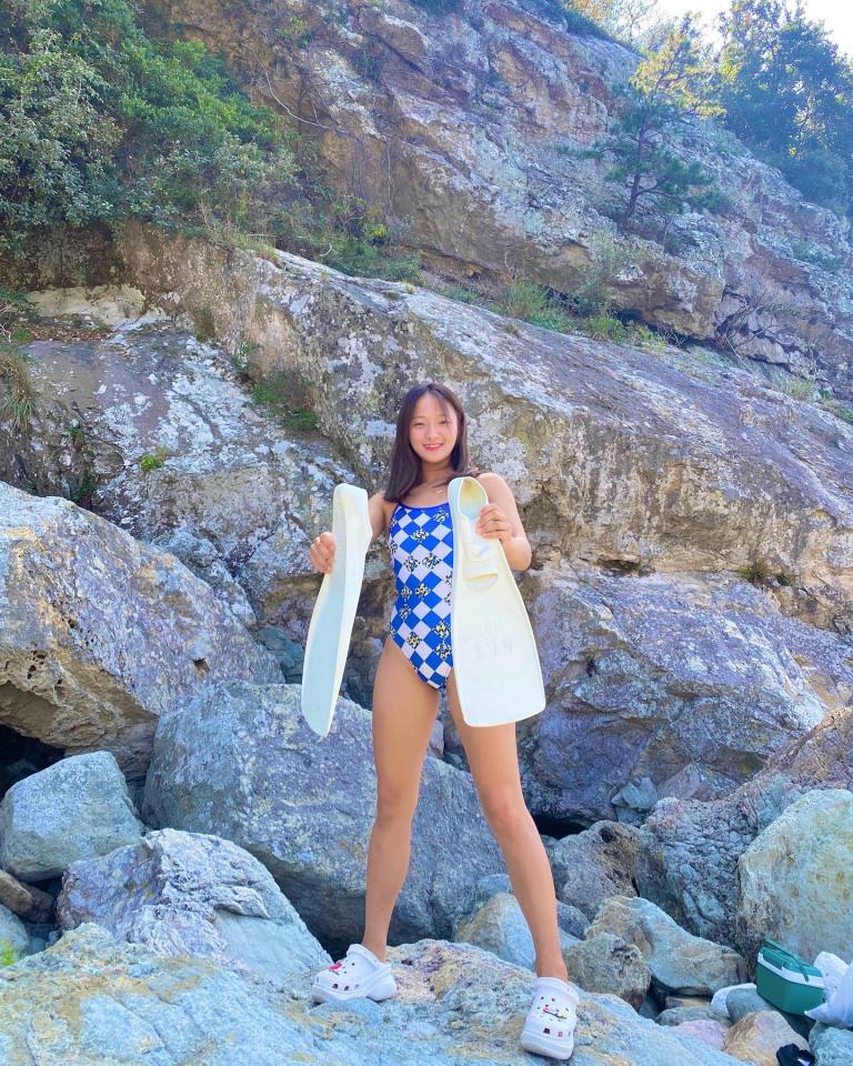 Woman in swimsuit holding swim fins by rocky shore.