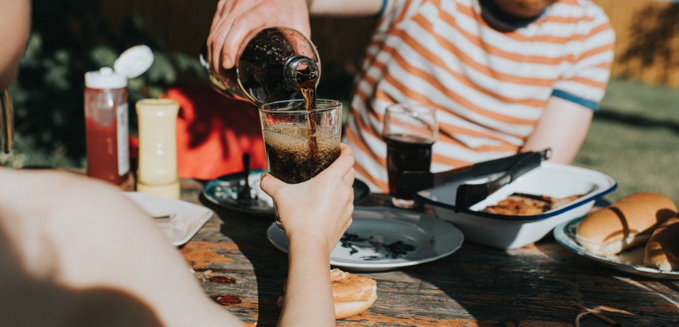 A child holds out a glass while an adult pours a fizzy drink at an outdoor barbecue.
