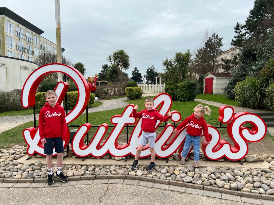 Three children in red Butlin's sweatshirts stand in front of a large Butlin's sign.