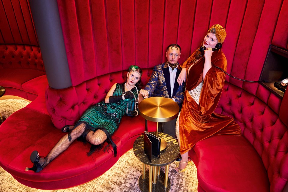 Three people in 1920s attire sitting on a red velvet couch.