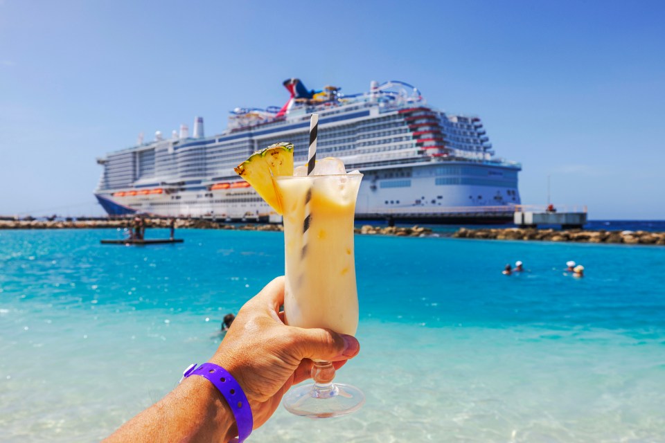 Hand holding a cocktail on a beach with a cruise ship in the background.