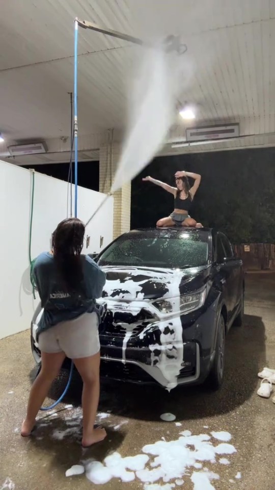 Two women washing a car at a car wash, one sitting on the roof.