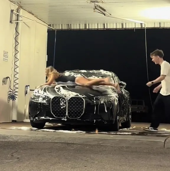 A woman lying on the hood of a car in a car wash, with a man watching.  The text overlay reads, "pov: the car wash trend needs a warning label."