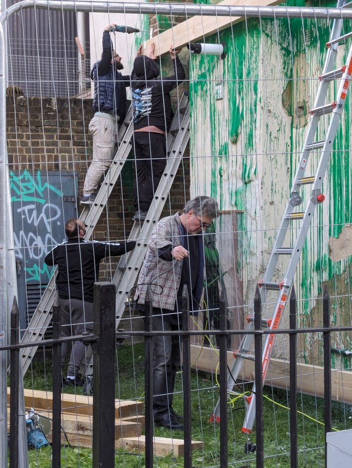 Workers installing a wooden beam near a green graffiti-covered wall.