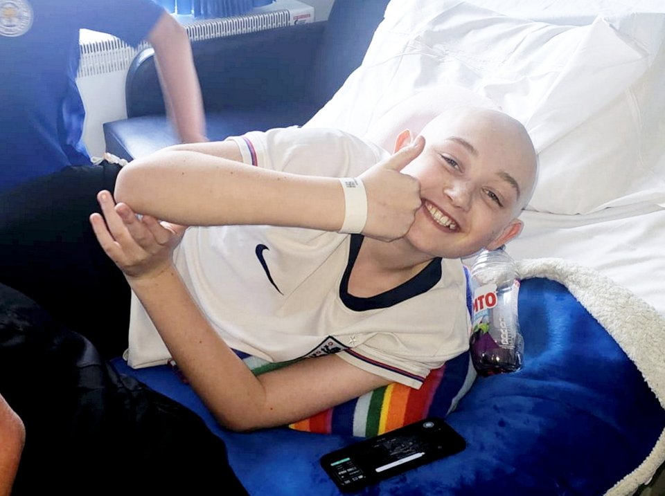 Smiling boy with shaved head giving thumbs up from hospital bed.
